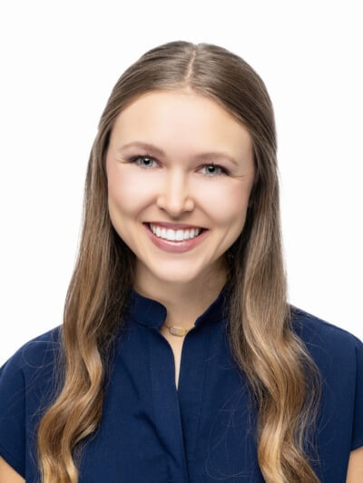A young woman with long, wavy light brown hair smiles at the camera. She is wearing a navy blue top and a delicate necklace. The background is plain white.