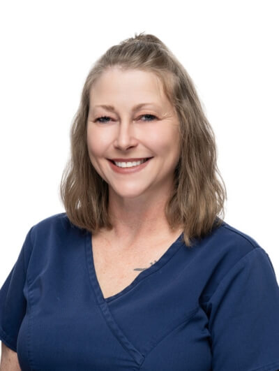 A woman with shoulder-length light brown hair, wearing a navy blue scrub top, smiles at the camera against a plain white background.