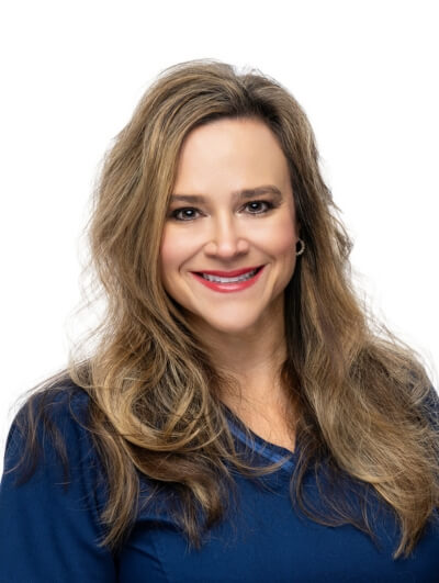 A woman with long, wavy light brown hair and fair skin smiles at the camera. She is wearing a navy blue top and has a neutral white background behind her.