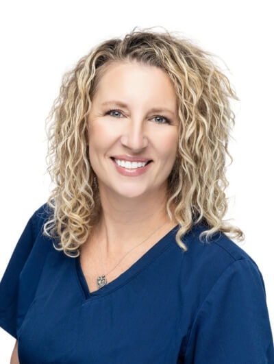 A woman with curly blonde hair, wearing a navy blue scrub top and a necklace, smiles at the camera against a plain white background.